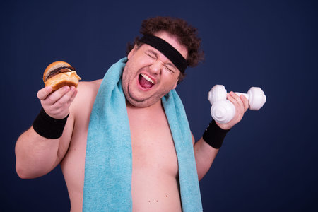 Sport, fitness and healthy lifestyle. Funny fat guy goes in for sports and eats a hamburger.