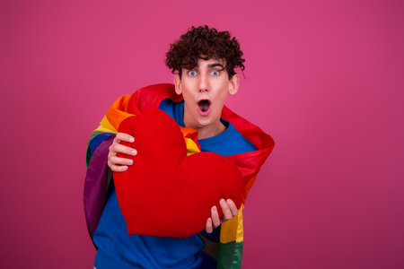 Surprised young man holding a heart shaped pillow and looking at camera on pink backgroundの写真素材