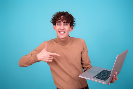 Portrait of a young man with curly hair in a beige sweater holding a laptop and pointing at it on a...