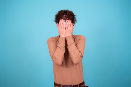 Portrait of a young man covering his face with his hands isolated on blue backgroundの写真素材