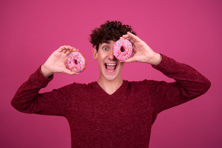 Portrait of young man with curly hair holding donuts isolated on pink background