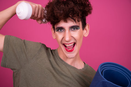 Cheerful young man holding yoga mat and shaker over pink background