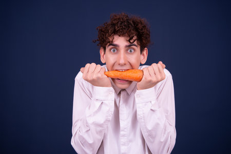 Young man with curly hair eating a carrot on a blue background.の写真素材