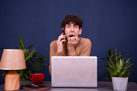 Portrait of a young man sitting at the desk with laptop and talking on the phoneの写真素材