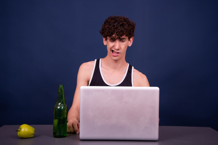 Young handsome man with curly hair sitting at the table with laptop and drinking beerの写真素材