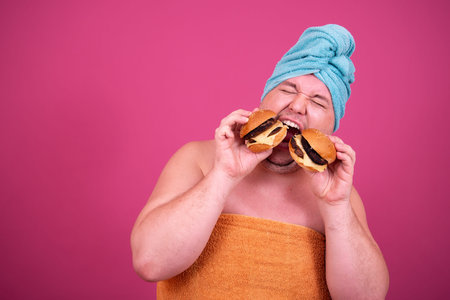 Fat man in orange towel and turban eating hamburger isolated on pink background