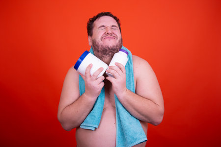 Fat man with a beard on a red background in a blue towel holds a bottle of protein powder in his handsの写真素材
