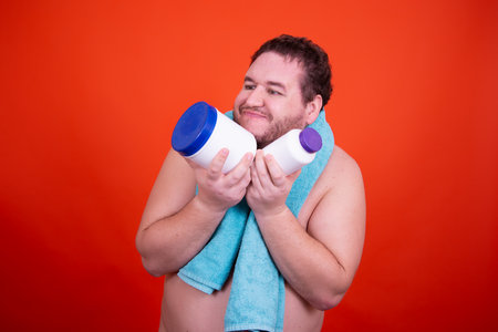 Portrait of a young man with a towel on his shoulders holding a bottle of protein powder in his hands on a red backgroundの写真素材