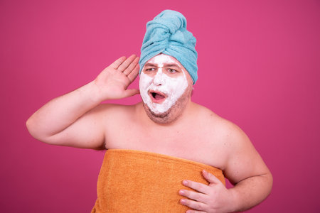 Portrait of a man with a towel on his head and a cosmetic mask on his face on a pink backgroundの写真素材