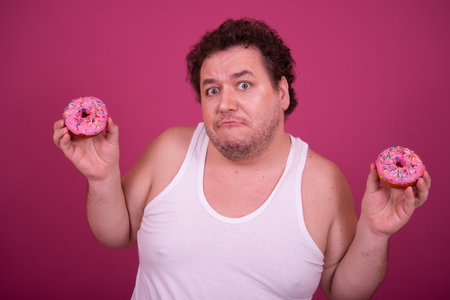 Fat man with curly hair holding donuts and looking at camera isolated on pink backgroundの写真素材