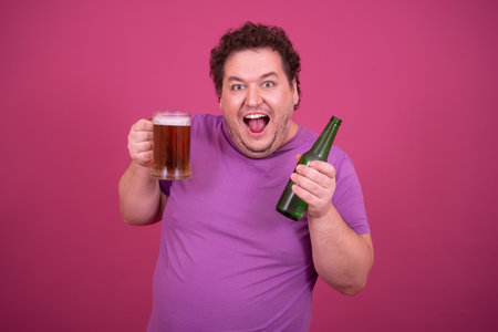 Portrait of happy overweight man smiling while drinking beer against pink backgroundの写真素材