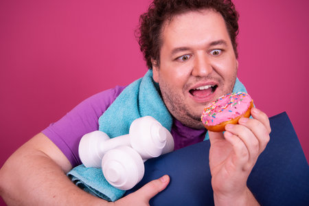 Fat man eating donut and dumbbells isolated on pink backgroundの写真素材