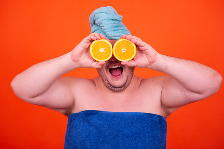 Surprised man with a towel on his head holding two halves of orange in front of his eyes