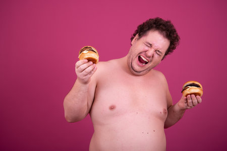 Fat man with curly hair eating hamburger isolated over pink background.の写真素材