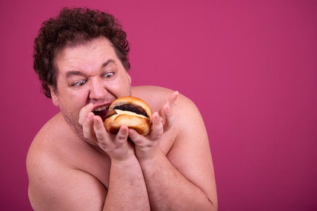 Portrait of fat overweight man with curly hair eating burger against pink backgroundの写真素材