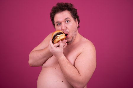 Fat overweight man with curly hair against pink background and eating hamburgerの写真素材