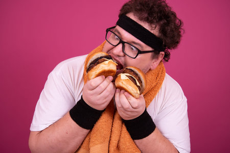 Funny fat man eating hamburger and looking at camera over pink background