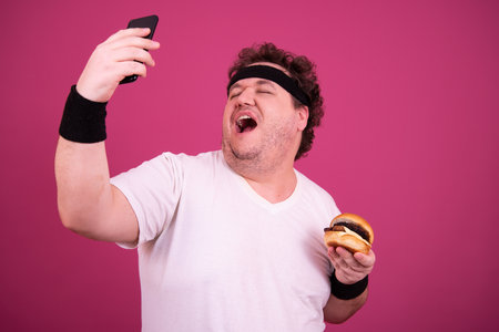 Funny fat man eating hamburger and taking selfie with smartphone isolated on pink backgroundの写真素材