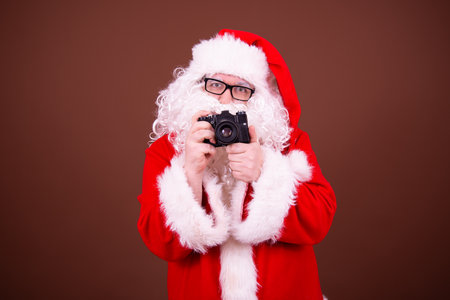 christmas, holidays, technology and people concept - man in costume of santa claus with camera over brown backgroundの写真素材