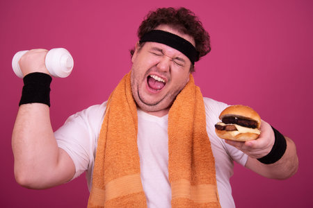 Fat man with dumbbell and burger isolated on pink background. Fat man in sportswear