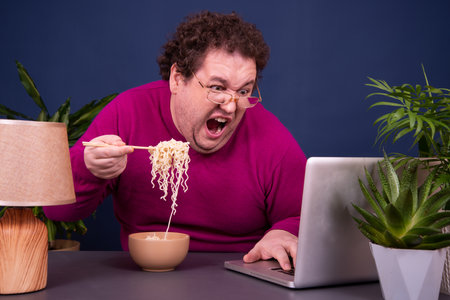 Funny fat man eating instant noodle while sitting at the table in front of laptopの写真素材