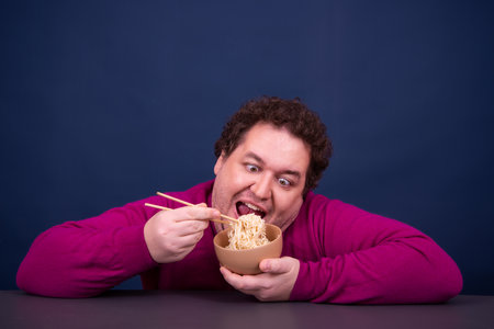 Funny fat man eating noodles with chopsticks, on blue backgroundの写真素材