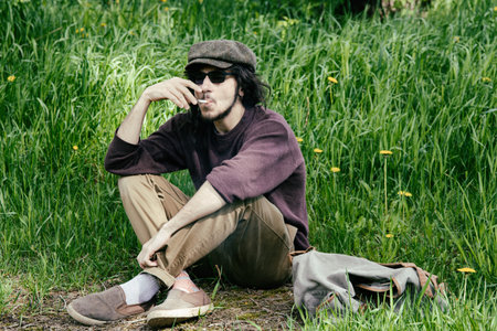 Young hipster man with hat and sunglasses sitting on the grass and talking on the phoneの写真素材