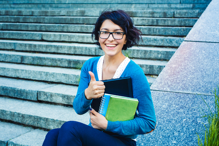 Student girl holding notebook and books. Young beautiful woman sitting on stairs and showing thumbs up.の写真素材