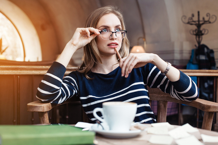 Serious young woman in glasses with wooden pencil in hand writing notes stickers in cafe with cup of coffee. Concept of planning personal traning schedule.Looking away.の写真素材