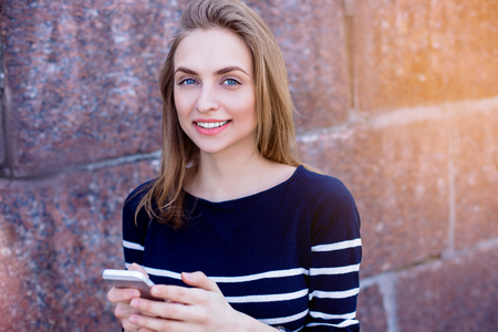 Beautiful young woman, blonde, making reminders outdoors using smartphone and fast 4G internet connection while standing against stone wall. Sunny spring day. Granite stone wallsの写真素材