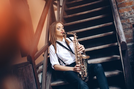Playing young attractive girl sitting on steps in white shirt with a saxophone - outdoor in old town. Sexy young woman with sax.の写真素材
