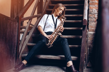 Beautiful young girl plays a saxophone sitting on steps - outdoors. Attractive woman in white shirt plays a saxophone.の写真素材
