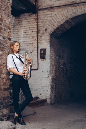 Beautiful young girl plays a saxophone standing near a white old wall - outdoors. Attractive woman in white shirt expression plays a saxophone.の写真素材