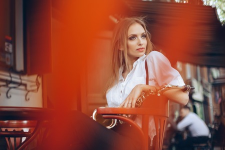 Young attractive girl in white shirt with a saxophone sitting near caffe shop - outdoor in sity. Sexy young woman with sax thinking about something.の写真素材