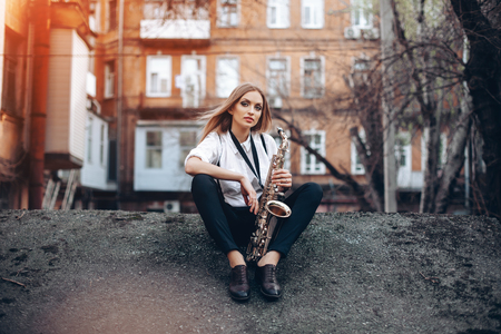 Young attractive girl in white shirt with a saxophone sitting sits on the earth - outdoor. Sexy young woman with sax looking at camera.の写真素材