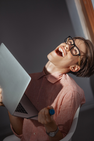 IT computer nerd. Creative teen millenial portrait indoors. Funny young man with happy face expression in glasses hug laptop against dark background. Internet, video game addiction conceptの写真素材