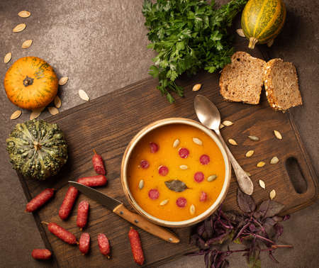 Pumpkin cream soup, served with smoked mini sausage and bread, parsley, basil, seeds on a cutter board. Stone background.の写真素材