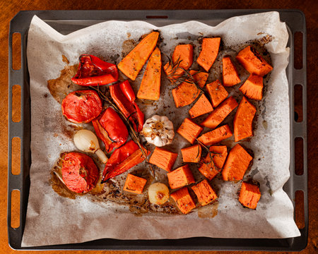 Roasted sweet potatoes, red bell peppers, tomatoes, garlic and onions on parchment paper baking tray, healthy autumn vegetable side dish, cozy home cooking, warm rustic kitchen moodの写真素材