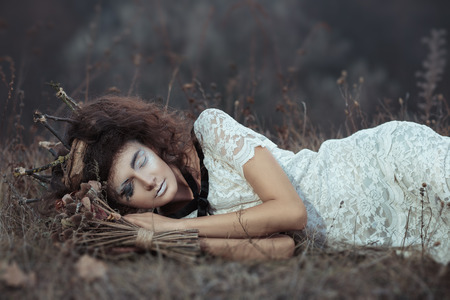 Young girl in the image of a bride in a white lace dress, a crown of twigs with a bouquet of dried flowers lying on the groundの写真素材