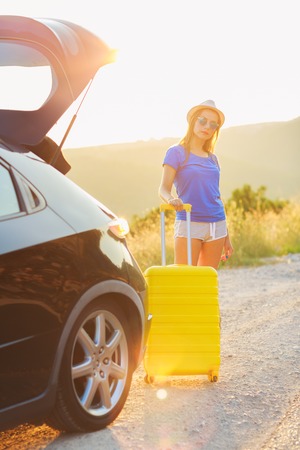 A young woman with a yellow suitcase standing near the trunk of a car parked on the roadsideの写真素材