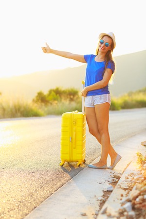 An attractive young woman with a yellow suitcase is traveling on the road hitchhikingの写真素材