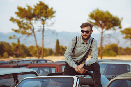 Portrait of a young handsome stylish man, wearing shirt and bow-tie on the field of old cars