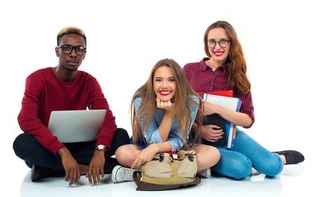 Three happy young teenager students sitting with books, laptop and bags isolated on white backgroundの写真素材