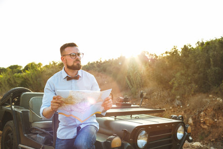Young stylish man with glasses and bow tie see the map near the old-fashioned SUV on the sunset