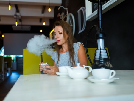 Beautiful young woman smoking a hookah and using smartphone in a cafeの写真素材