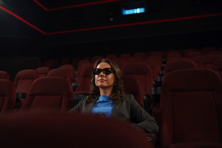 Young woman watches movie 3D at the cinema and drinks coffee, looking at the screen, alone in an empty room, overall planの写真素材