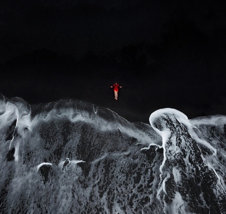 Aerial view of a girl in a red dress sitting on the beach with black sand, foaming waves of the Atlantic Ocean. Tenerife, Canary Islands, Spainの写真素材