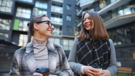 Two happy women walking with takeaway coffee and talking with interest among themselves in the business district.の写真素材