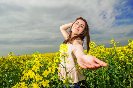 Young woman standing in yellow rapeseed field smiling at camera  With one hand in her hair and one...