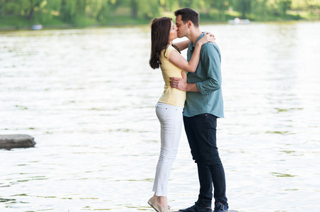 Joyful and spontaneous couple in love, holding hands on a stone pier on a natural lake having fun, kissing, hugging and laughing outdoorsの写真素材